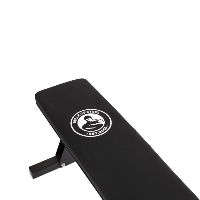 The Flat Utility Bench from Bells of Steel features a sturdy design with a robust metal frame and a sleek black finish. It proudly displays the "Bells of Steel" logo, established in 2010, which depicts a muscular arm flexing inside a circle, signifying its maximum capacity for serious weight training.