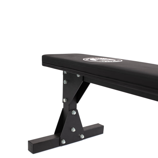 Introducing the Bells of Steel Flat Utility Bench, designed in black with a robust structure featuring metal supports and secure bolts. The padded surface proudly displays the brand logo. This bench is equipped with a cross-shaped base to provide enhanced stability, making it perfect for strength training exercises. With its impressive max capacity, it's built to endure intense workouts. The product is showcased against a plain white background.