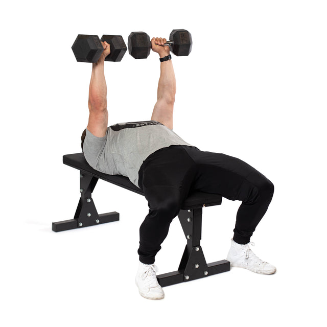 A person wearing a gray t-shirt, black pants, and white shoes performs a dumbbell bench press on the Bells of Steel Flat Utility Bench. They confidently extend their arms straight up while holding a dumbbell in each hand, demonstrating the impressive max capacity of the bench's sturdy design.