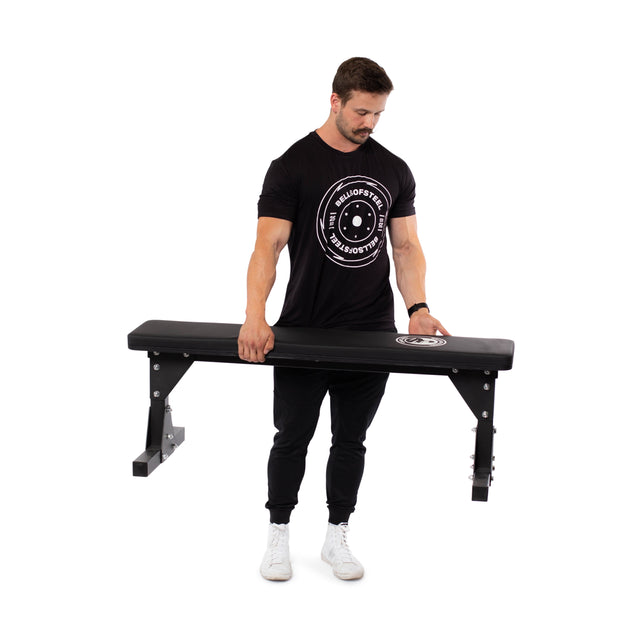 A person dressed in a black t-shirt and black pants is holding a Bells of Steel Flat Utility Bench, featuring a logo. The bench's sturdy design ensures maximum support, while the person is positioned against a white background.