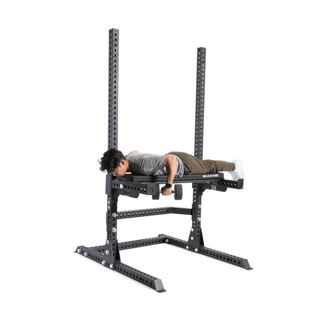 A person is lying face down on a reverse hyperextension machine, gripping the handles near the 3"x3" rack. This Bells of Steel Canada product, specifically the Utility Seat for Hydra/Manticore with its black metal frame and adjustable components, is situated in a well-lit room that also features a versatile exercise bench.