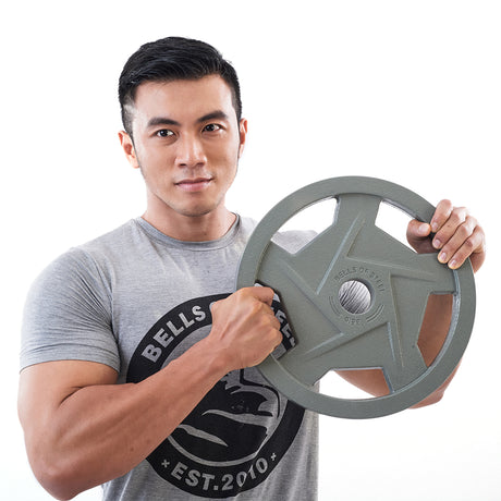 A man in a gray T-shirt, displaying the "Bells of Steel" brand and "Est. 2010," confidently holds a large Gray Mighty Grip Olympic Weight Plate with both hands. Standing against a plain white background, he gazes directly at the camera, demonstrating the formidable hold of his Bells of Steel Olympic training gear.