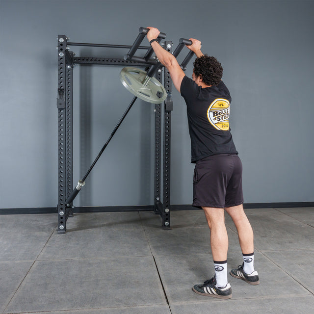 Wearing a black shirt and shorts, a person stands on the gym floor holding the handles of a Bells of Steel Viking Press Attachment fixed to an Olympic bar with weights, getting ready to work out.