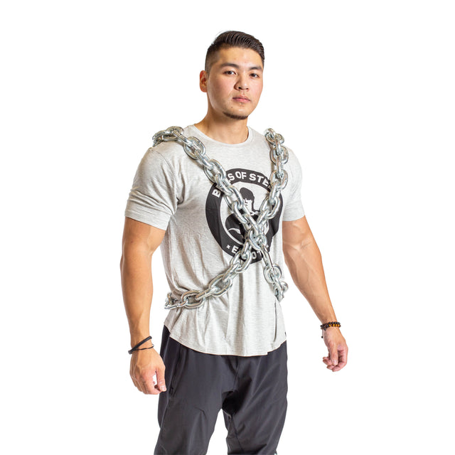 A man stands confidently in a Bells of Steel Chain Weight Vest, with weighted chains forming an X across his gray t-shirt and black pants. The background is white.