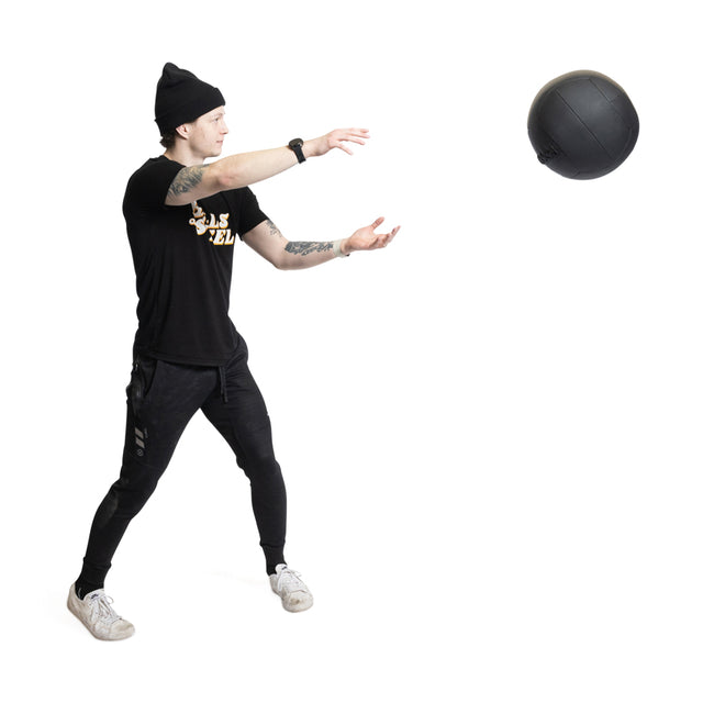 A person in a black beanie, T-shirt, and pants showcases diverse fitness levels by tossing the Bells of Steel's Triple Stitched Medicine Ball. They highlight this exercise with tattoos on one arm against a stark white background.