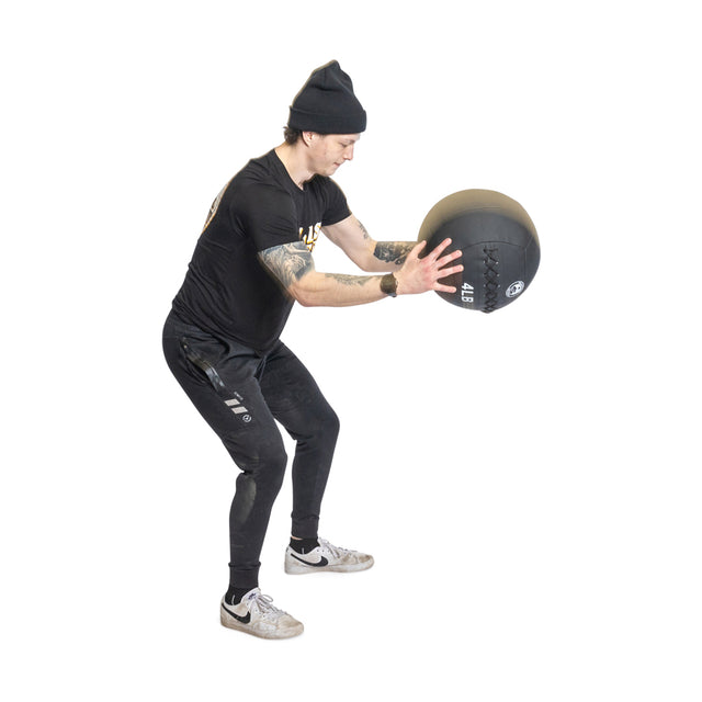 Someone in black athletic clothing and a beanie holds a Bells of Steel Triple Stitched Medicine Ball with a Kevlar outer shell. They stand in an athletic stance, leaning slightly forward, and appear focused against the plain white background.