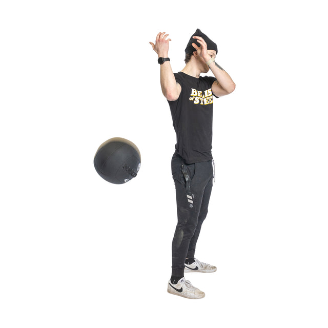 A person in casual athletic wear, featuring a black beanie and Nike sneakers, holds their head with one hand while a large Bells of Steel Triple Stitched Medicine Ball seems to float nearby. Their shirt is adorned with yellow text.