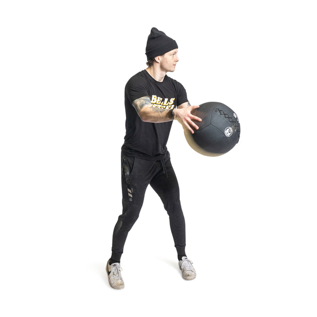 In a gym setting, an individual wearing a beanie, black t-shirt, and joggers is holding the Triple Stitched Medicine Ball from Bells of Steel.