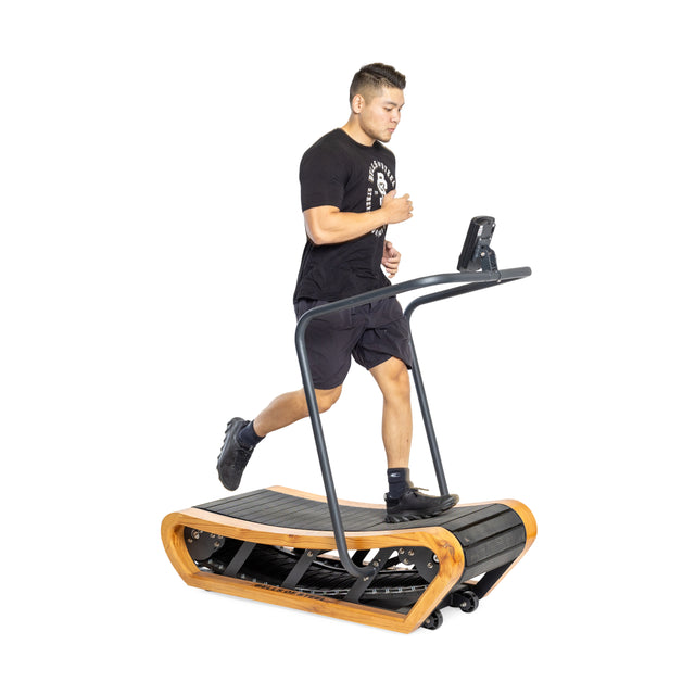 A person runs on the Bells of Steel Wooden Residential Manual Treadmill, featuring impact-absorbing slats, a wooden frame, and a black belt. The small display complements their sleek black athletic gear against a plain white background.