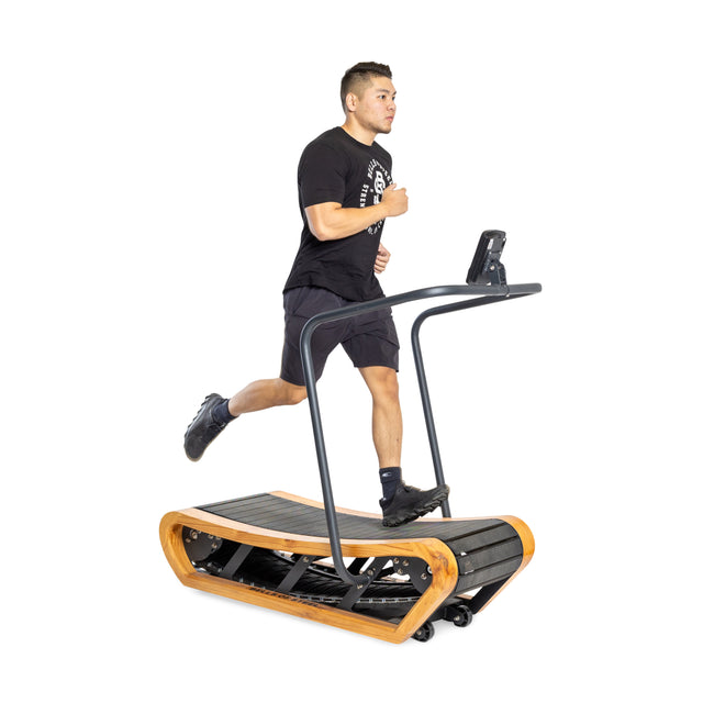 A man dressed in black is jogging on the Bells of Steel Wooden Residential Manual Treadmill, featuring high-strength nylon slats, a wooden frame, and a digital display set against a plain white background.