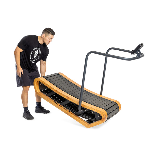 Someone wearing a black shirt and shorts adjusts a Bells of Steel Wooden Residential Manual Treadmill with a sleek black belt, wooden frame, simple metal handlebar, and small console on a plain white background.