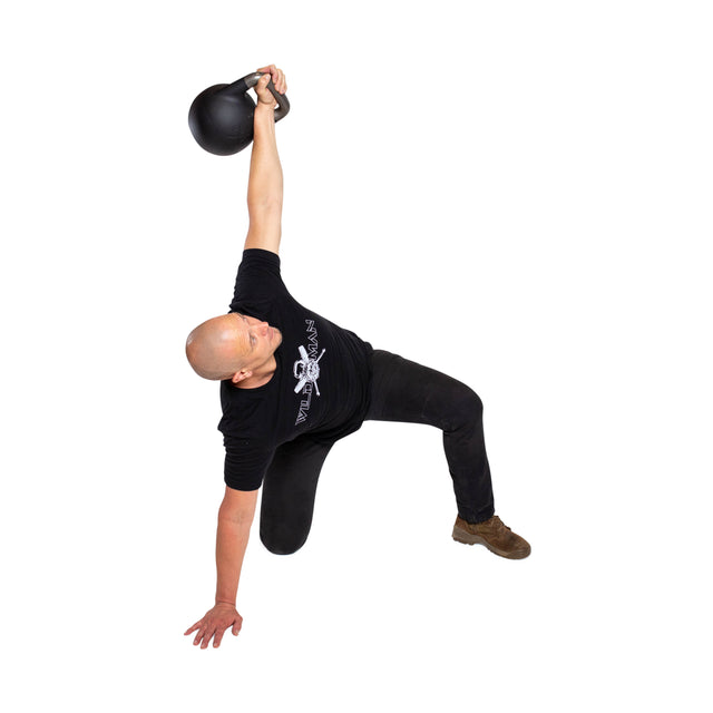 A person in a black T-shirt, black pants, and brown shoes performs a Turkish get-up with one arm holding a Mark Wildman Adjustable Kettlebell by Bells of Steel, kneeling against a white background.