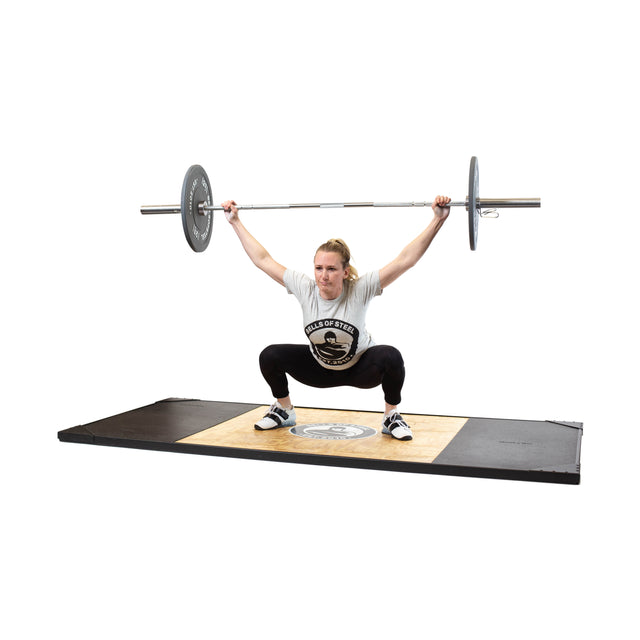 Wearing a gray T-shirt, black pants, and weightlifting shoes, a person demonstrates Olympic lifting precision performing an overhead squat on a platform with the Bells of Steel Oly Lifting Needle Bearing Bar - Women's.