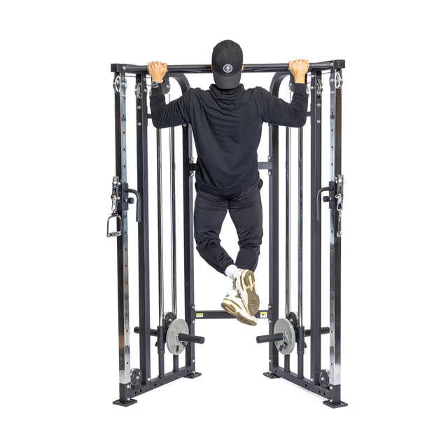 A person in a black hoodie, pants, and cap is doing pull-ups on the Bells of Steel Functional Trainer. This machine features weight stacks and steel bars for cable exercises, against a plain white background emphasizing strength training.