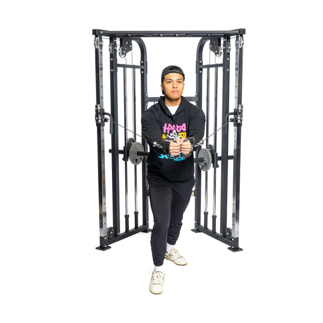 A person intensely uses the Bells of Steel Functional Trainer, pulling cables forward in a staggered stance while wearing black pants, a hoodie, and sneakers, embodying focus and strength through functional training.