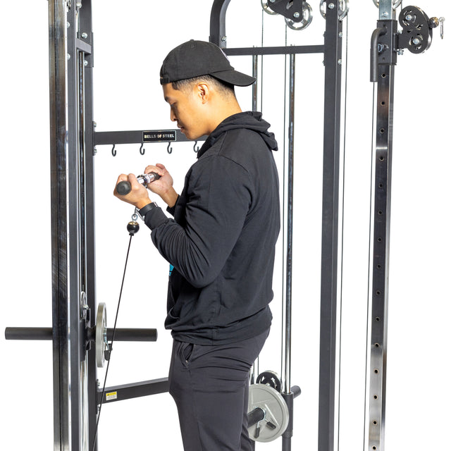 A man in black athletic wear and a cap performs cable curls using the Bells of Steel Functional Trainer. The machine, featuring weight stacks, allows him to focus on his exercise against the plain white background.