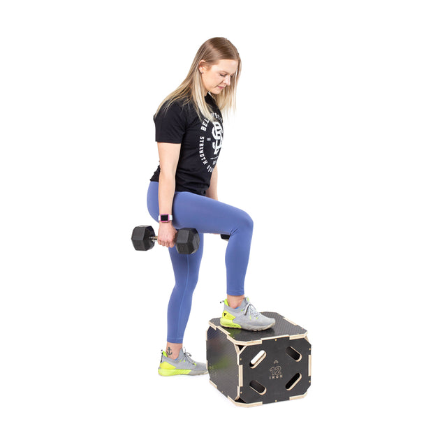 A woman works out using a Bells of Steel 3 in 1 Anti-Slip Wood Plyo Box, stepping onto the platform while holding a dumbbell in one hand. She's dressed in a black shirt, blue leggings, and gray sneakers with bright accents. The plyo box is labeled with "12.2 inches" and provides three height options for diverse exercises.