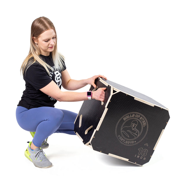 A person wearing a black shirt and blue leggings is kneeling and adjusting a Bells of Steel "3 in 1 Anti-Slip Wood Plyo Box" labeled "18 inch." This versatile home gym equipment showcases a sleek logo and provides three different heights in one, making it perfect for varied workouts.