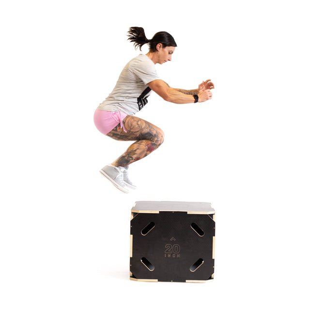 A tattooed individual is captured mid-jump in a gym setting, sporting a grey t-shirt and pink shorts. They are performing a box jump onto the Bells of Steel 3 in 1 Anti-Slip Wood Plyo Boxes. This versatile home gym equipment provides three adjustable heights, ideal for boosting your workout. The background is plain white.