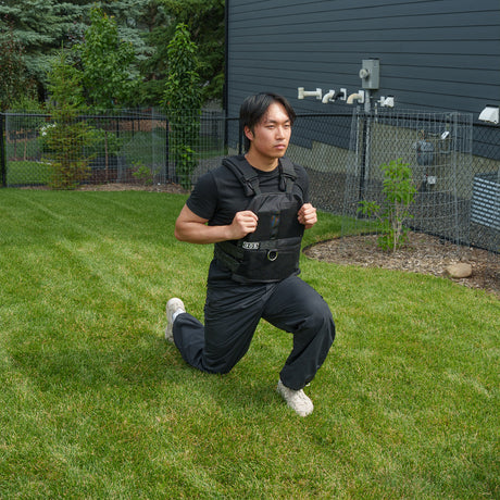 Wearing the Bells of Steel Ultimate Backyard Training Bundle, a person dressed in black workout clothes performs lunges on a grassy lawn in their fenced backyard, maximizing their outdoor workout with this essential fitness equipment.
