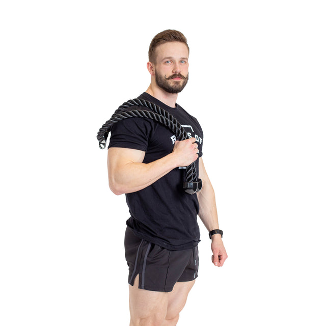 A man with a beard is holding the Bells of Steel Extra Long Tricep Rope Extension (137cm) over his shoulder, prepared for some intense exercises. He’s wearing a black t-shirt and shorts against a crisp white background.