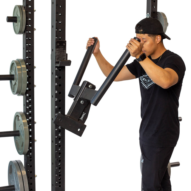 Clad in a black outfit, an individual adjusts the Bells of Steel Y Dip Bar Rack Attachment on a power rack, with an assortment of barbell plates visible in the background. The gym setting showcases equipment designed for strength training.