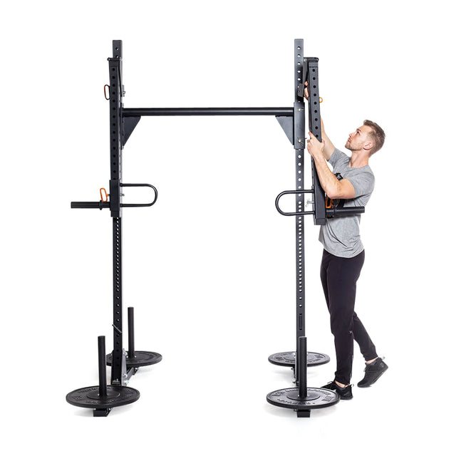 A man in athletic clothing adjusts the height of a pull-up bar on a Bells of Steel Strongman Yoke, which doubles as a power rack. The yoke features several weight plates and pegs for stability. The background is white.