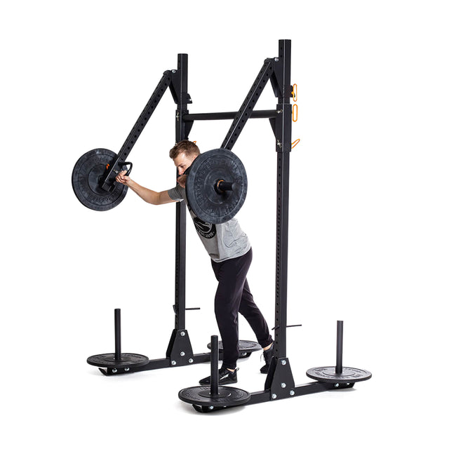 A person wearing a gray shirt and black pants works out with a landmine attachment on a Bells of Steel Strongman Yoke. They are pushing a weighted barbell connected to the rack, which features organized weight plates on pegs and safety bars for secure lifting.