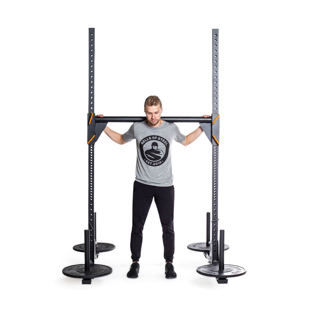 An individual wearing a gray T-shirt and black pants is lifting the Bells of Steel Strongman Yoke in a gym, with weight plates loaded onto it. The yoke, which features orange accents and adjustable ⅝" holes on its 2.3" x 2.3" frame reminiscent of a squat stand, serves as the focal point as they concentrate intensely on the exercise.