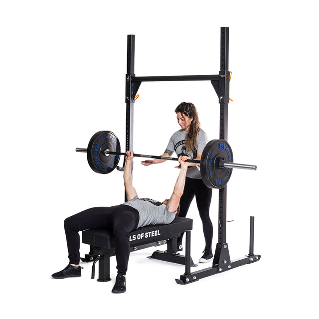 A person is lying on a bench lifting weights in a bench press, assisted by another person. They are using the Bells of Steel Strongman Yoke, featuring 2.3" x 2.3" dimensions and ⅝" holes for weight pegs, and both are dressed in athletic apparel. The background is plain white.