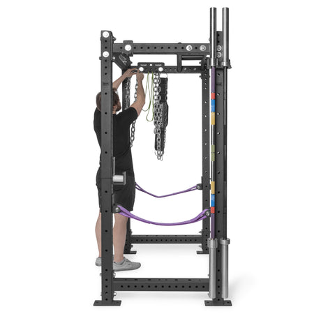 A person adjusts chains and resistance bands on a power rack. Nearby, a Bells of Steel Accessory Holder (Wall or Rack Mounted) organizes weightlifting bars and barbell collars as a convenient gym storage solution.