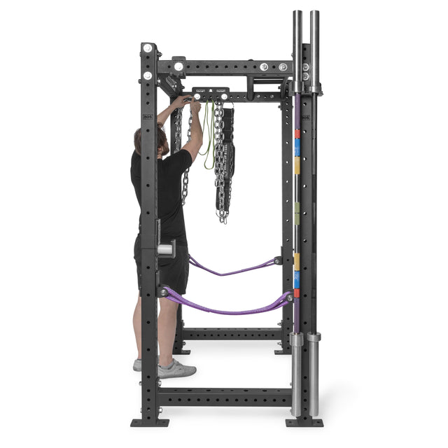 A person adjusts chains and resistance bands on a power rack. Nearby, a Bells of Steel Accessory Holder (Wall or Rack Mounted) organizes weightlifting bars and barbell collars as a convenient gym storage solution.