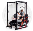 A man aids a woman in lifting the Home Gym Builder barbell, enhanced with resistance bands for bench pressing within power racks. Both are wearing gray shirts adorned with a black Bells of Steel logo, set against a white backdrop featuring a semi-circular dotted design.