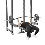 A person in black athletic clothing and a cap is lying on a "Bells of Steel" weight bench, lifting a heavy-duty barbell. The home gym setup includes the Spotter Arms Rack Attachment with UHMW lining for added safety.