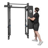 A man in sportswear attaches the Bells of Steel Wall or Rack Mounted Bench Hanger to a black metal power rack designed for strength training and gym storage, set against a plain white background.