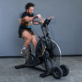 A man pedals fast on the Bells of Steel Blitz Max Air Bike with fan resistance, causing motion blur in his arms and legs. The air bike has extra wheels on its front base. The background is a plain gray wall.