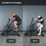 Two people, one 6'2" and one 5'1", ride the Bells of Steel Blitz Max Air Bike side by side in a gym. Text at the top reads: "Suitable for all heights.