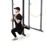 Dressed in black, a person uses the Bells of Steel Split Squat Leg Roller Rack Attachment for stability while performing a Bulgarian split squat with a dumbbell. One leg rests on the rack amidst a minimal white background with yellow band accents.