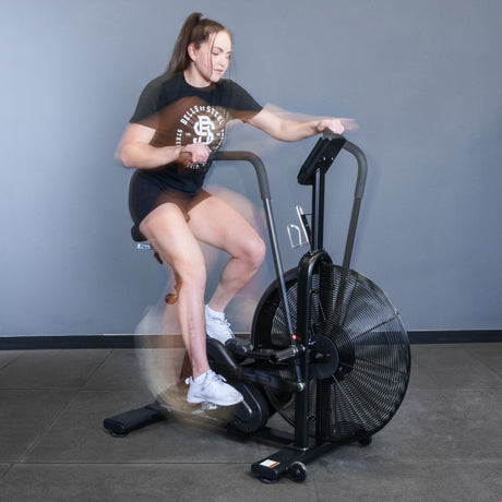 A woman in athletic wear works out on the Bells of Steel Blitz Air Bike against a gray wall. Her blurred motion highlights intense pedaling and arm movement on this commercial-grade air bike.
