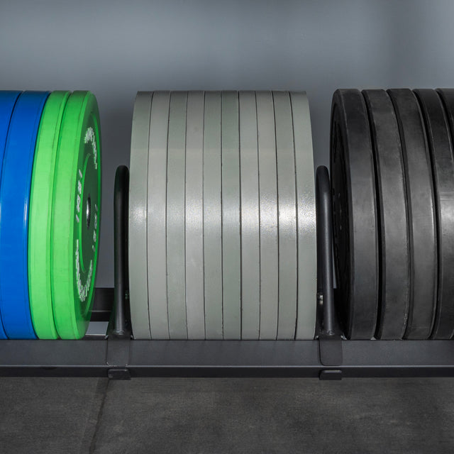 The Bells of Steel Divider for Bumper Plate Shelf (Pack of 3) keeps green, blue, gray, and black weight plates neatly organized on a gym floor against a gray wall.