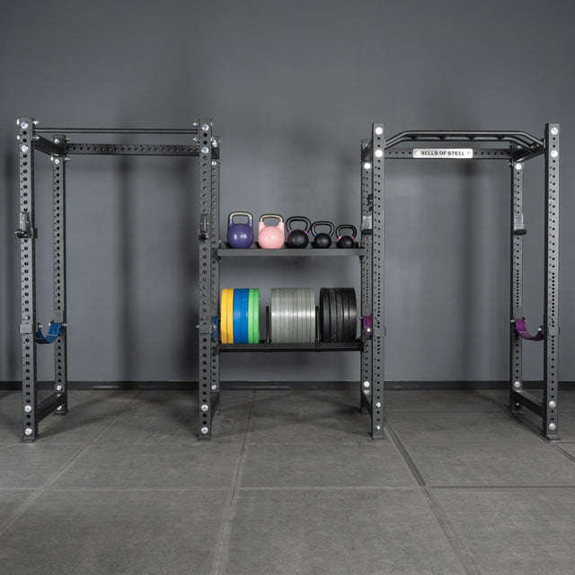 A gym setup with metal squat racks, colorful Bells of Steel kettlebells, and weight plates neatly organized on storage shelves using Bells of Steel Divider for Bumper Plate Shelf (Pack of 3), all set against a gray wall and floor.