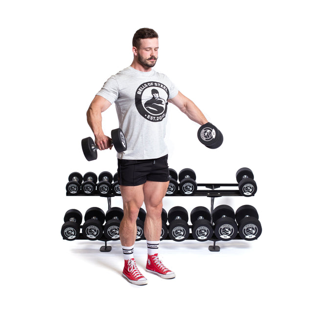 A man in a grey t-shirt and red sneakers does a lateral raise with dumbbells, standing before the Bells of Steel Commercial Interchangeable Weight Rack on a white background.