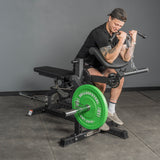 A man in a black shirt, shorts, and backward cap uses the Bells of Steel Legacy Leg Extension / Hamstring Curl Machine - Plate Loaded at home, gripping the handles and flexing his arms.