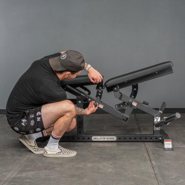 A person kneels beside the Bells of Steel Legacy Leg Extension / Hamstring Curl Machine - Plate Loaded in a home gym, adjusting its angle by hand; the Bells of Steel branding is visible on the machine.