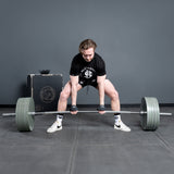 A man in athletic wear lifts a heavily loaded Bells of Steel Deadlift Bar, displaying powerlifting strength on a gray gym floor with a black box and gym logo in the background.