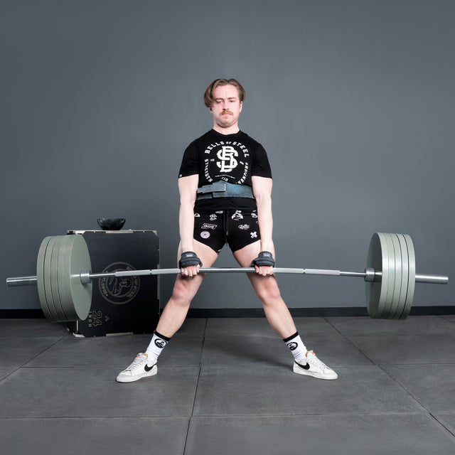 A person wearing a black t-shirt, shorts, and white sneakers demonstrates powerlifting strength by performing a sumo deadlift with a heavily loaded Bells of Steel Deadlift Bar in a gym with gray walls and floor mats.