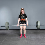 A person with long hair, glasses, and pink socks is powerlifting in a gym using the Bells of Steel Deadlift Bar, standing on a grey floor against a plain grey wall.