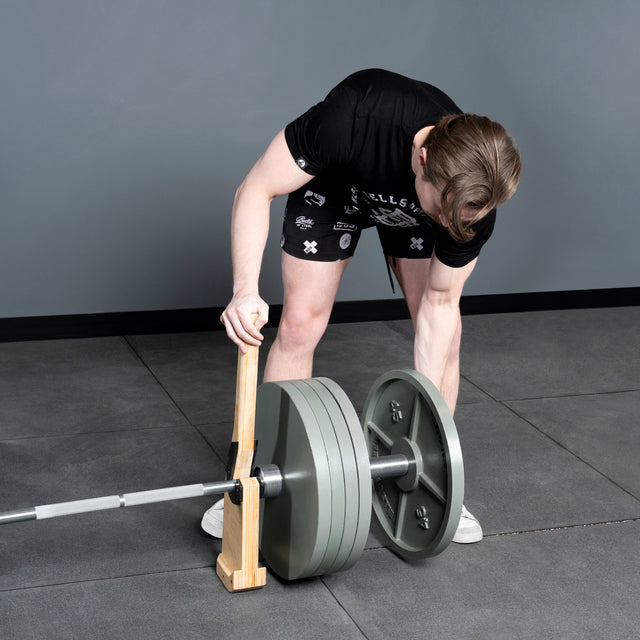 A person in a black shirt and shorts uses a wooden lever to load heavy weight plates from the Bells of Steel Deadlift Bar on a gym floor with gray walls, demonstrating powerlifting and strongman techniques.