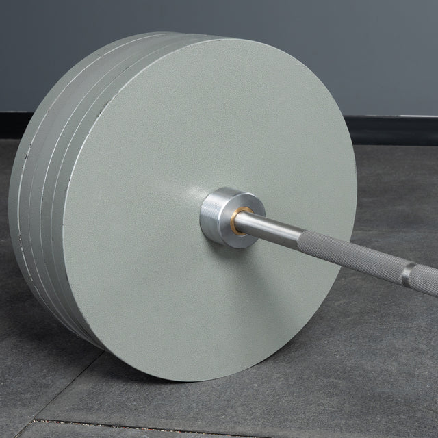 A Bells of Steel Deadlift Bar loaded with large, light gray plates rests on a gym floor, set against a gray wall.