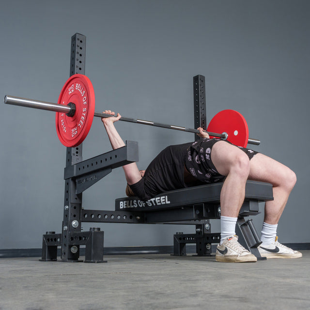 Wearing athletic gear, a person lies on the Bells of Steel Hydra Two Post Converter Bench, gripping a barbell with red plates as they get ready to perform a bench press in the gym.